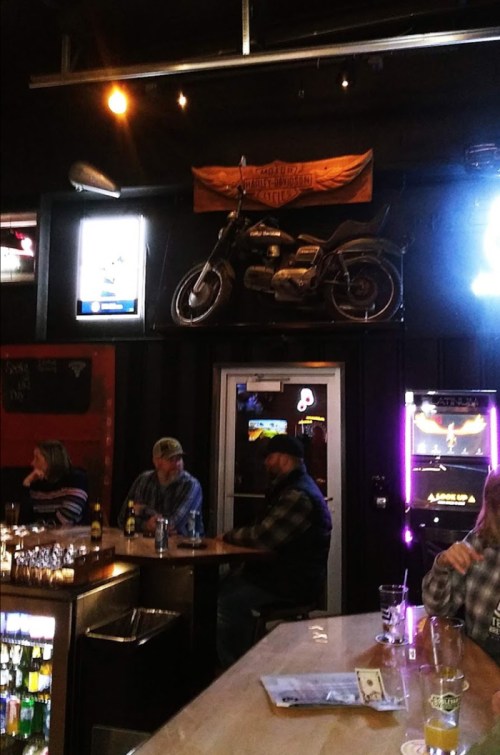 A bar scene with patrons, a motorcycle on the wall, and a brightly lit jukebox in the background.