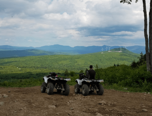 Two people sit on ATVs overlooking a lush green valley with wind turbines in the distance under a cloudy sky.