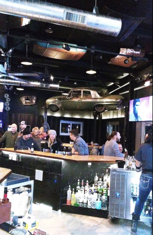 A lively bar scene with patrons at the counter, a vintage car hanging from the ceiling, and a TV displaying a performance.
