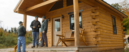 Three men stand on the porch of a log cabin, engaged in conversation, surrounded by trees and autumn foliage.