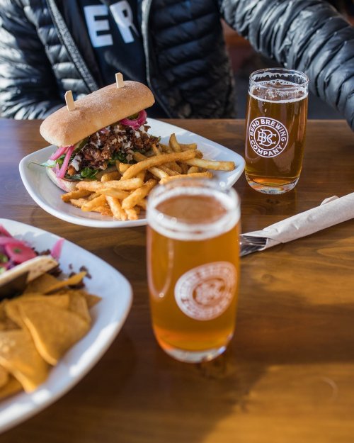 A plate with a sandwich, fries, and two glasses of beer on a wooden table. A person in a black jacket is nearby.