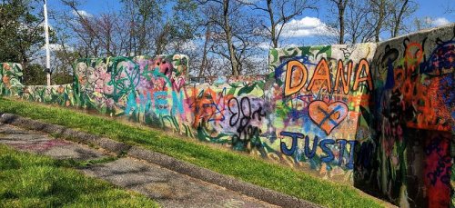 Colorful graffiti covers a concrete wall, featuring names and hearts, with trees and a blue sky in the background.