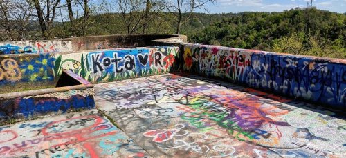 A colorful, graffiti-covered concrete platform surrounded by trees and hills under a clear blue sky.