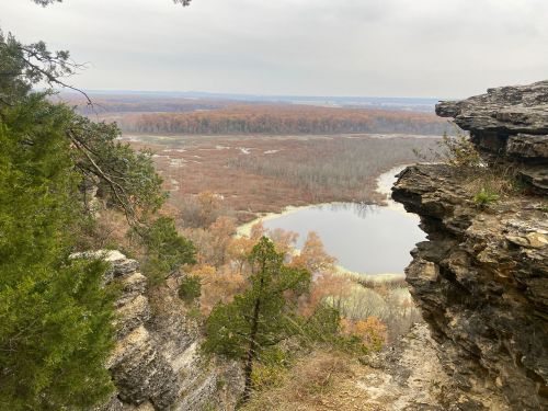 A scenic view from a rocky cliff overlooking a calm pond surrounded by autumn foliage and distant hills.