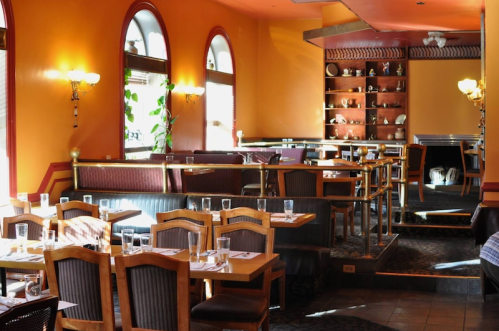 Interior of a cozy restaurant with wooden tables, chairs, and warm orange walls, featuring soft lighting and decorative shelves.