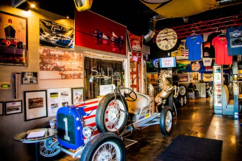 A vintage race car with an American flag design inside a museum, surrounded by racing memorabilia and posters.