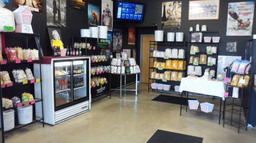 Interior of a snack shop with shelves of popcorn and drinks, movie posters on the walls, and a cooler in the corner.