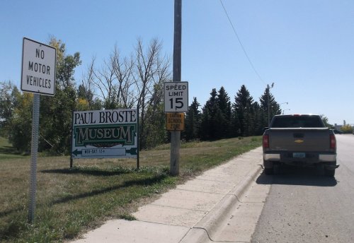 A sign for Paul Broste Museum next to a road with a speed limit sign and a no motor vehicles sign.