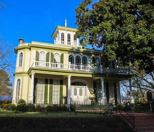 A large, historic yellow house with a balcony, surrounded by greenery and a clear blue sky.