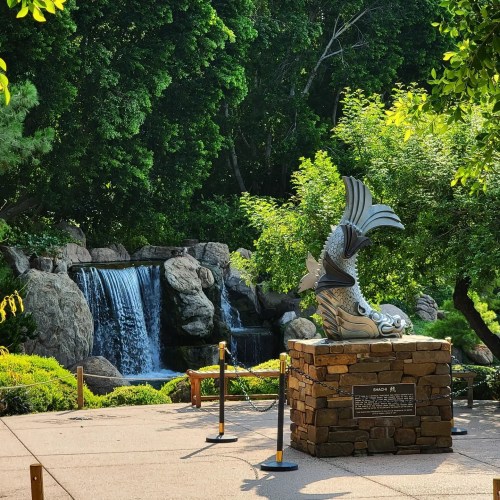 A serene garden scene featuring a waterfall and a decorative stone statue of a fish surrounded by lush greenery.