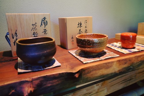 Three traditional Japanese tea bowls displayed on a wooden shelf, accompanied by decorative wooden boxes.