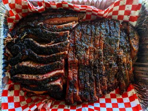 Sliced smoked brisket on a bed of red and white checkered paper in a foil tray.