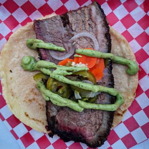 A close-up of a taco with brisket, sliced onions, jalapeños, carrots, and green sauce on a soft tortilla.