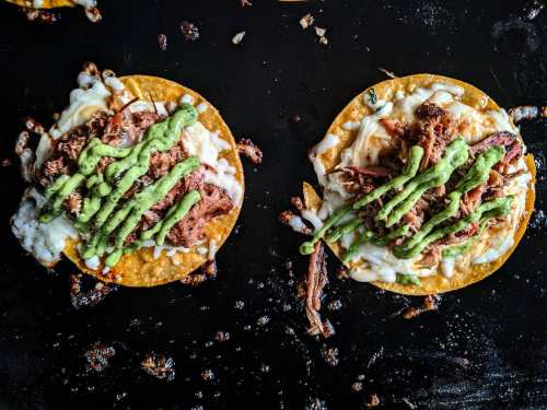 Two tacos on a black surface, topped with shredded meat, cheese, and green sauce, garnished with herbs.