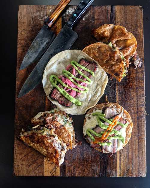 A wooden cutting board with four tacos topped with various sauces, alongside two sharp knives.