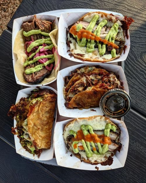 A variety of tacos and burritos topped with green sauce and salsa, served in white containers on a black table.