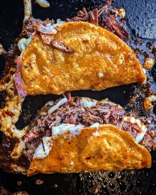 Two crispy tacos filled with shredded beef and melted cheese, sizzling in a pan with a golden-brown exterior.