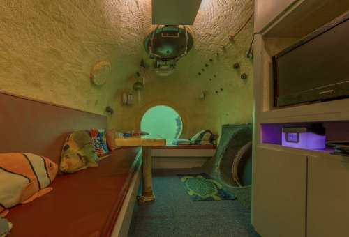 Interior of an underwater habitat with cozy seating, colorful decor, and a view of the ocean through a large window.
