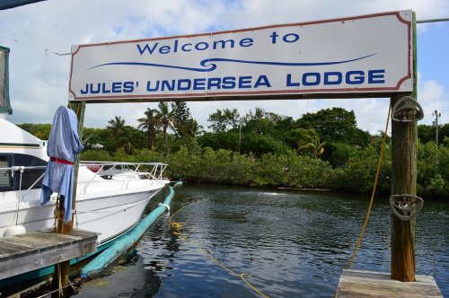 Sign reading "Welcome to Jules' Undersea Lodge" near boats and lush greenery in a tropical setting.