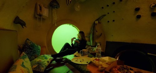 A person sits in a cozy, underwater-themed room, enjoying food with a large circular window showing green water outside.