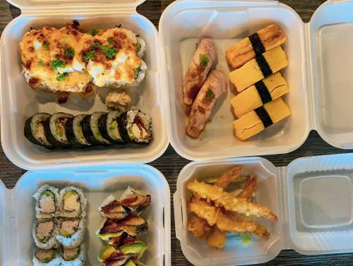 A variety of sushi and Japanese dishes in takeout containers, including rolls, tamagoyaki, and tempura.