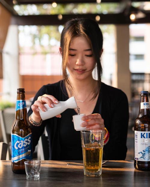 A woman pours a drink from a small bottle into a cup, with two beer bottles on the table beside her.
