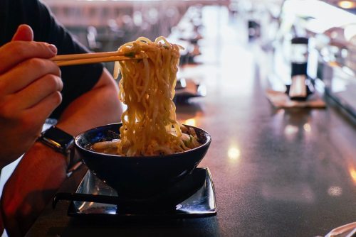 A person holding chopsticks with noodles above a bowl of ramen on a restaurant table.
