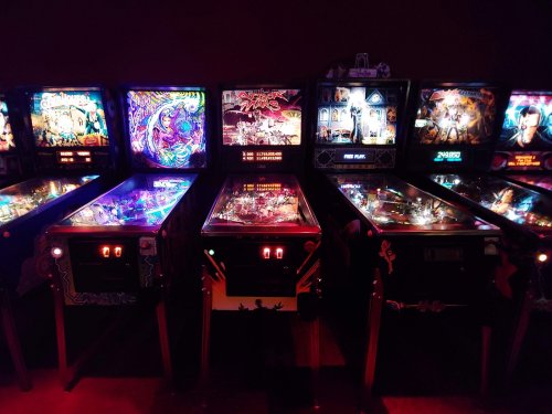 A row of colorful pinball machines illuminated in a dimly lit arcade.