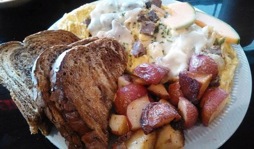A plate of breakfast featuring an omelet with meat, toast, and roasted potatoes, garnished with melon slices.