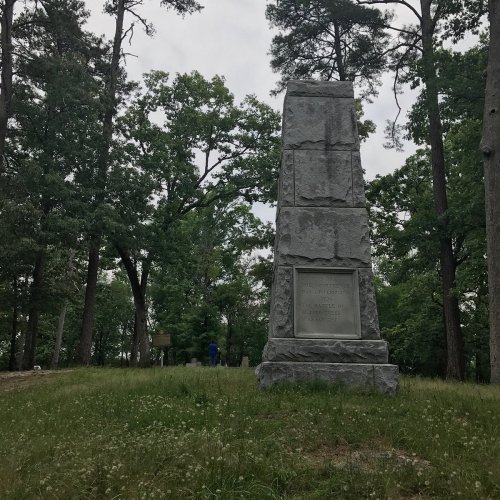 A tall stone monument surrounded by trees, with a grassy area in the foreground and a person in the background.