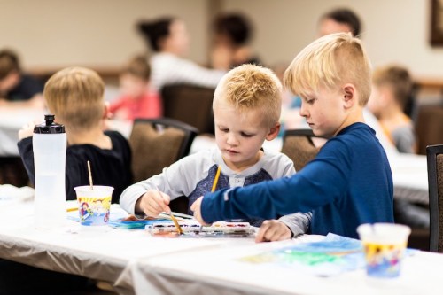 Two young boys are painting at a table, surrounded by other children in a classroom setting.