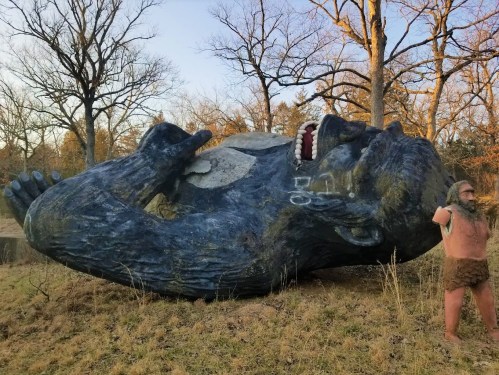 A large, blue gorilla statue lies on its back, with a smaller figure of a caveman standing beside it in a natural setting.