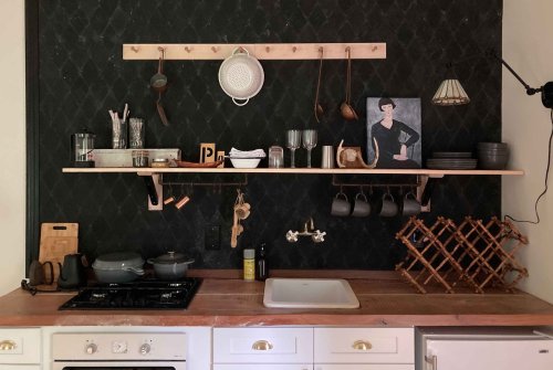 A stylish kitchen with dark walls, wooden shelves, cookware, glassware, and a framed portrait on display.