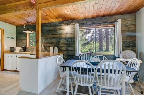 Cozy dining area with a wooden table and chairs, featuring a large window overlooking trees and a rustic kitchen.