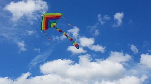 A colorful rainbow kite soaring in a bright blue sky with fluffy white clouds.