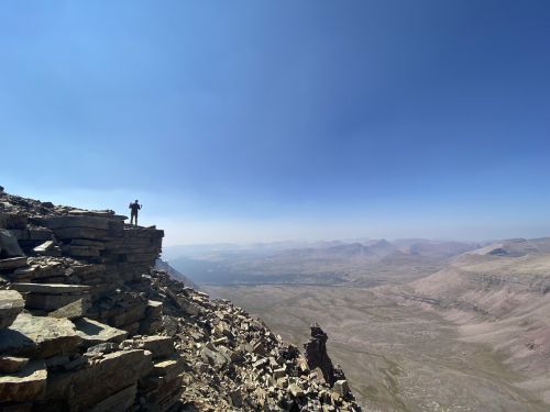 A hiker stands on a rocky mountain ledge, overlooking a vast, arid landscape under a clear blue sky.