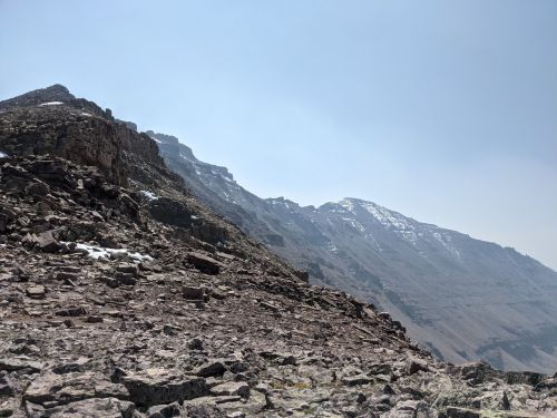 Rocky mountain landscape with steep slopes and a hazy sky, featuring patches of snow on the peaks.