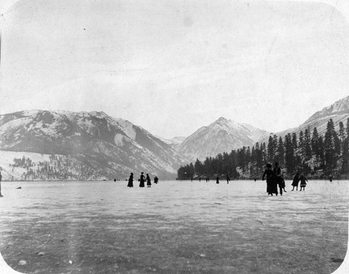 Black and white photo of people ice skating on a frozen lake, surrounded by snow-covered mountains and trees.