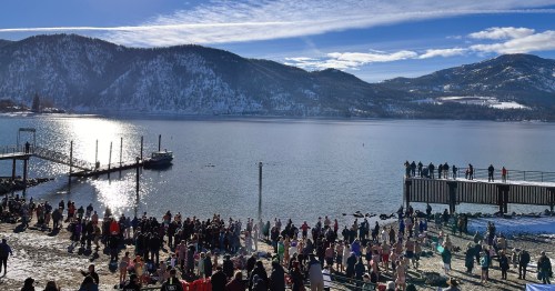 A crowd gathers by a snowy lake with mountains in the background, enjoying a sunny winter day.