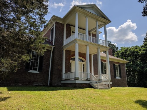 A historic brick house with white columns and a porch, surrounded by trees and a grassy lawn under a blue sky.