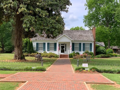 A charming blue house with white trim, surrounded by greenery and a brick pathway, featuring American flags on the porch.