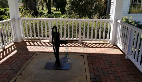 A vintage hand pump stands on a brick patio, surrounded by greenery and a white railing. Sunlight casts shadows.