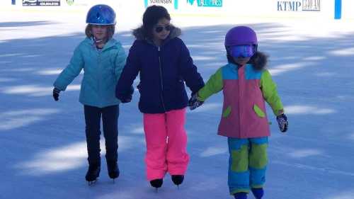 Three children ice skating hand in hand, wearing colorful winter jackets and helmets on an outdoor rink.