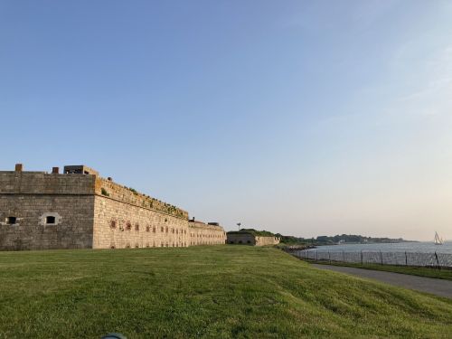A historic stone fort with grassy grounds, overlooking a calm waterway under a clear blue sky.