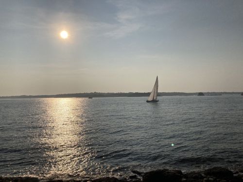 A sailboat glides across a calm sea under a hazy sun, with distant land visible on the horizon.