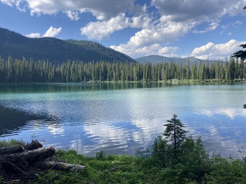 A serene lake surrounded by lush green forests and mountains, reflecting clouds in the clear blue water.