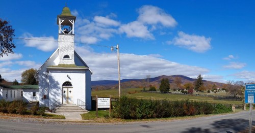 A white church with a bell tower, surrounded by green fields and mountains under a blue sky with clouds.