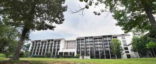 A modern multi-story building surrounded by trees and greenery under a cloudy sky.