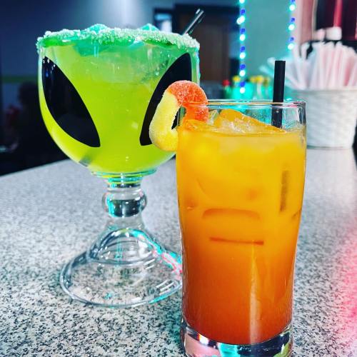 Two colorful cocktails on a bar: a green alien-themed drink and a vibrant orange beverage with a gummy garnish.