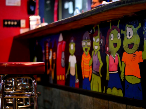 Colorful mural of cartoonish green characters on a bar counter, with a metal stool in the foreground.